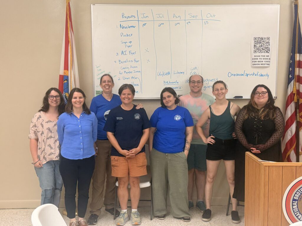 Photo of in-person attendees of Education committee meeting in May. Folks are standing by the whiteboard and flags in the union hall meeting space. In the picture, from left to right: Jade Ehrenzeller, Megan Kuhn, Amy Schwarzer, Windy Wood, April Brauda, Corey Kirk, Jaime Suarez Roy, Alena Santos.